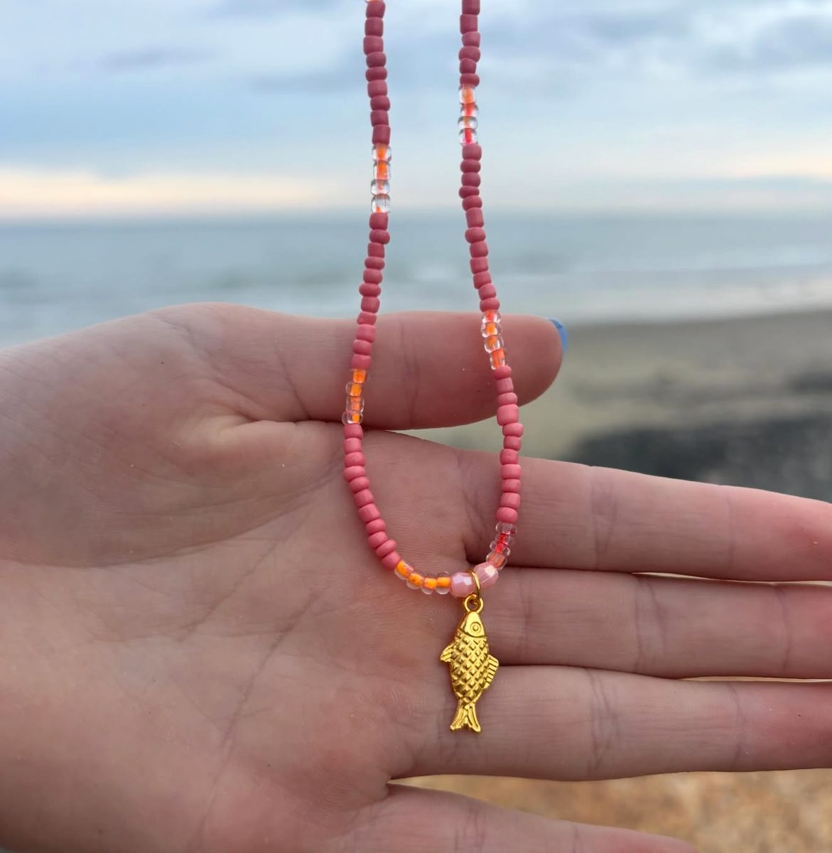 Pink beaded piece with a gold fish charm held in an open hand with the ocean in the background