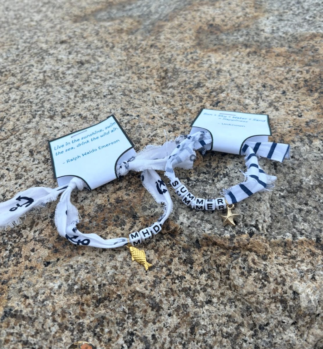 Two white fabric bracelets with letter beads and gold charms displayed on Marblehead rocks with quote cards behind them